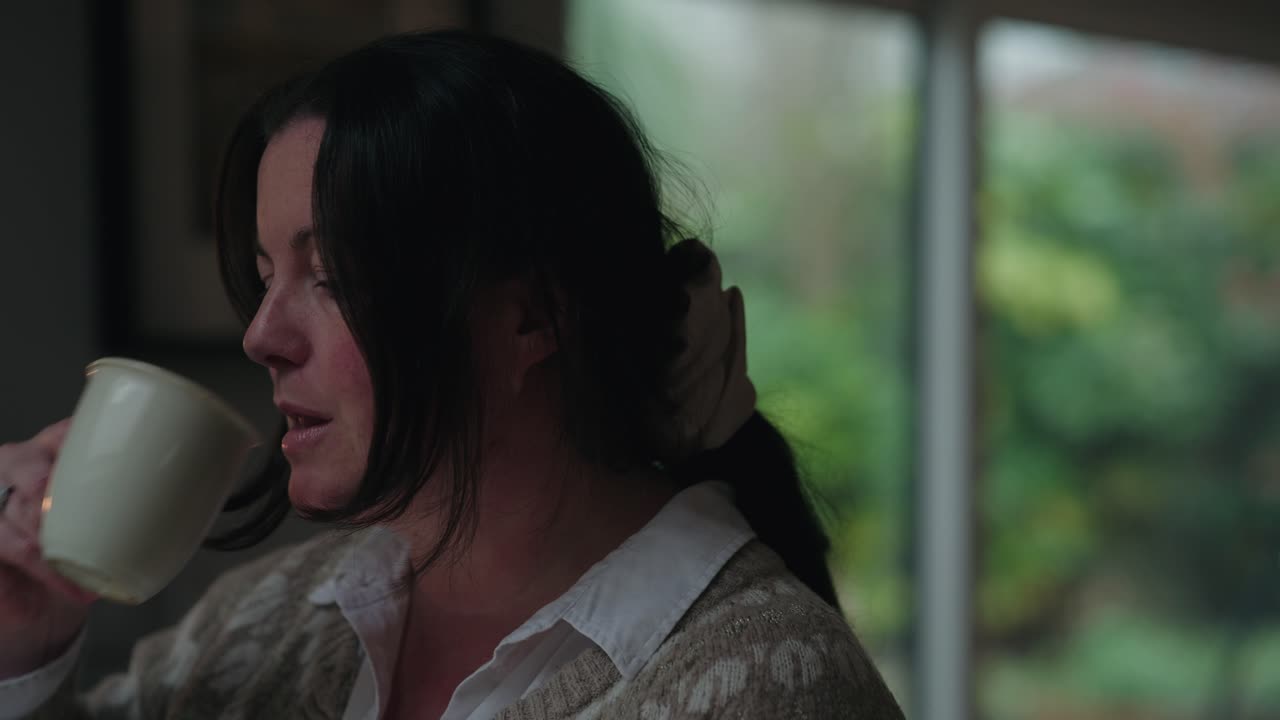 A close-up of a middle-aged woman with a focused expression, working at home near a bright window. Soft natural light and blurred greenery create a peaceful atmosphere.