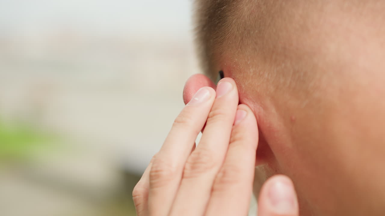 primer plano de un hombre quitándose un tapón del oído en un balcón, con la nuca sudada y el pelo corto visible. Los dedos pellizcan suavemente el tapón mientras las gafas de sol se apoyan en la montura de la oreja. Se aprecia un ligero enrojecimiento y la textura de la piel, lento y cuidadoso.