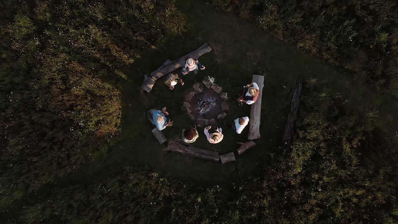 Drone Shot Of Mature Friends Making Toast As They Sit Around Fire At Outdoor Campsite