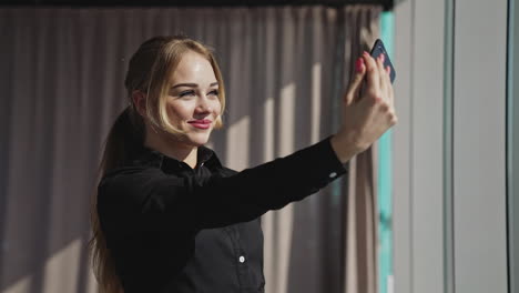 smiling attractive young adult woman holding phone and making selfie. happy beautiful lady professional smile stand posing alone indoors near the window in the sun.