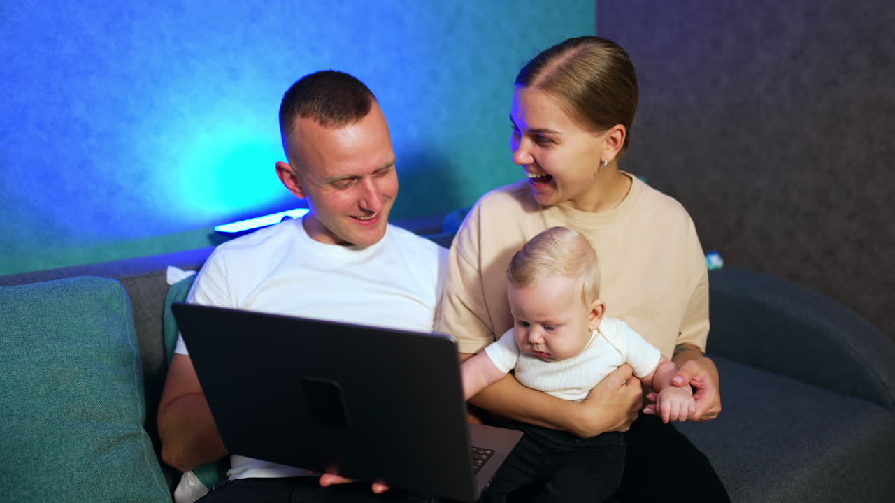 Happy smiling Caucasian people look at laptop. Little infant sitting in mom's hands is focused on computer screen.