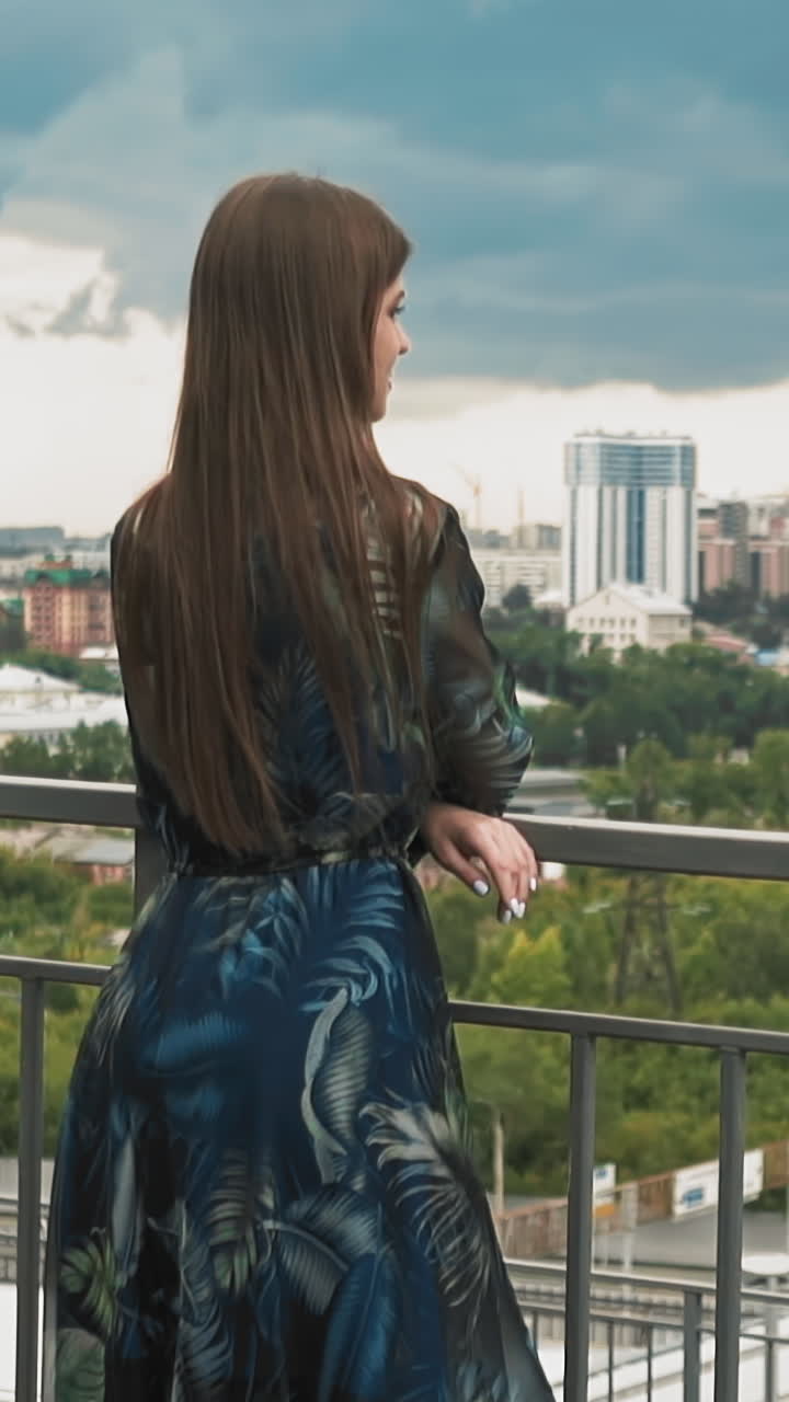 Thoughtful lady with loose hair wearing gorgeous chiffon dress leans on handrail on viewing deck against large city under cloudy sky slow motion backside view