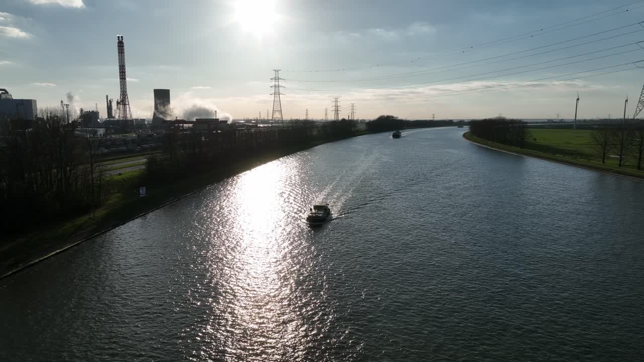 Zoom out aerial view of a boat navigating Antwerp industrial canal with factories, chimneys, and power lines under afternoon sun