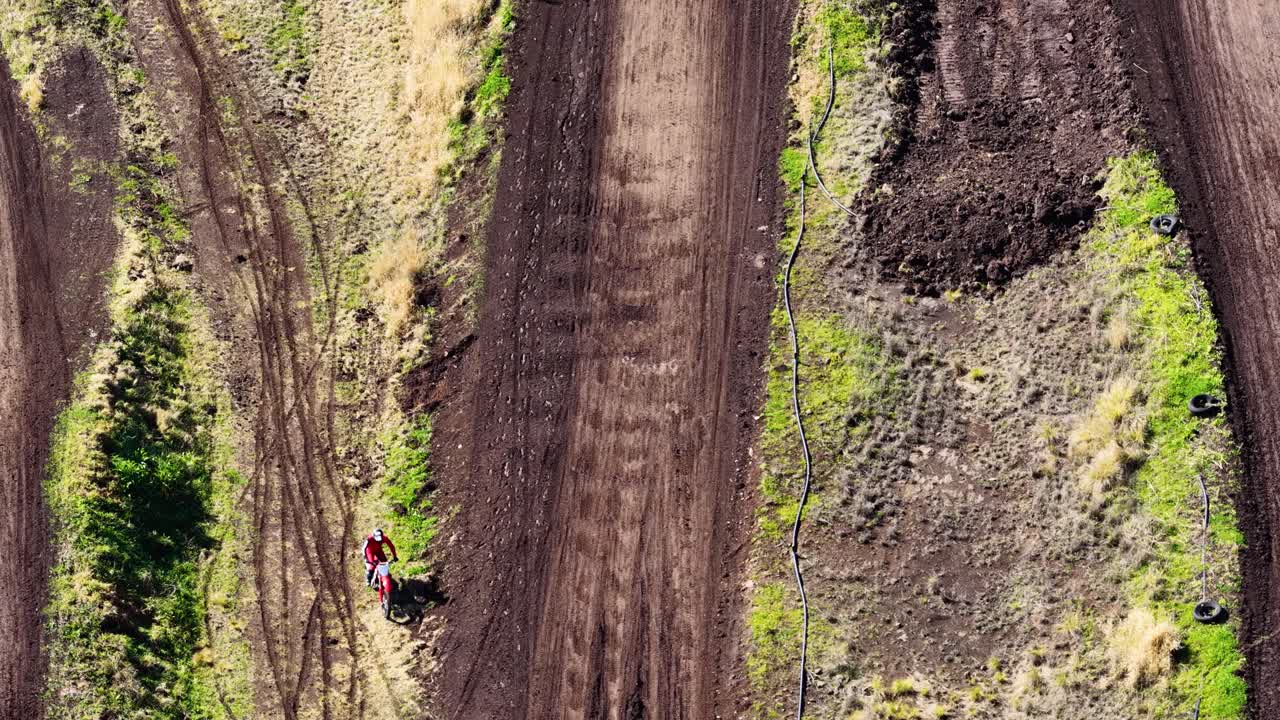 A motocross rider on a dirt bike jumps over a dirt track in a rural, bushland setting. Captured in bright daylight with aerial drone footage
