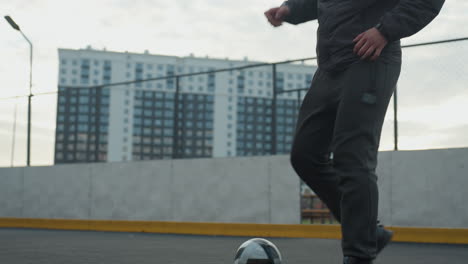 man skillfully controlling soccer ball with precise footwork on sport arena during intense training session, urban background features tall residential buildings, fencing, and goalpost