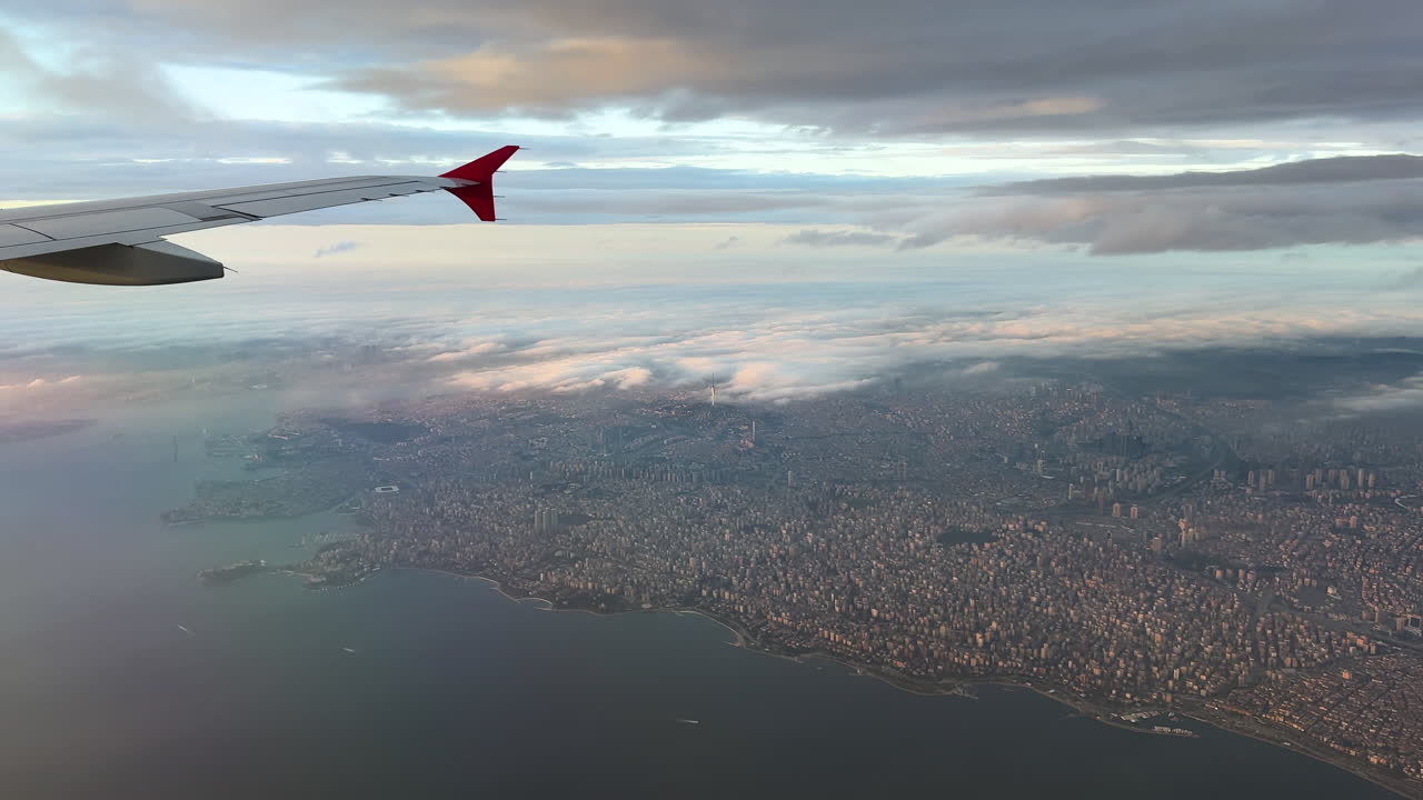 Luftfoto fra et kommercielt fly, der viser vingen over Istanbul med en bred kystbyudsigt nedenunder. Tæt bylandskab møder havet, delvist dækket af bløde skyer og varmt horisontlys.