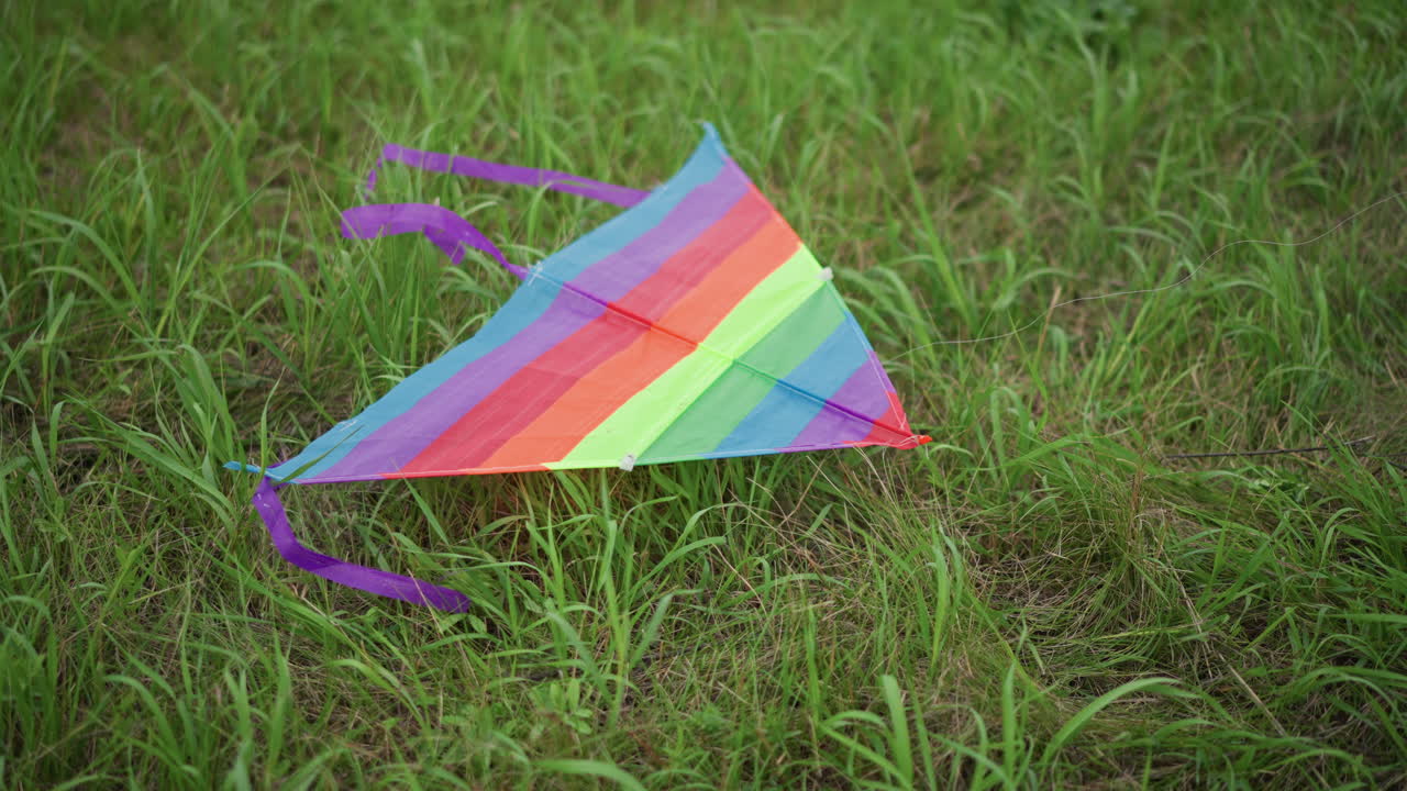 Un colorato aquilone adagiato sull'erba, tessuto arcobaleno a righe con coda viola poggiato su un prato baciato dalla rugiada, un primo piano cinematografico suggestivo che evoca la nostalgia dell'infanzia e una malinconica atmosfera estiva, bokeh delicato.