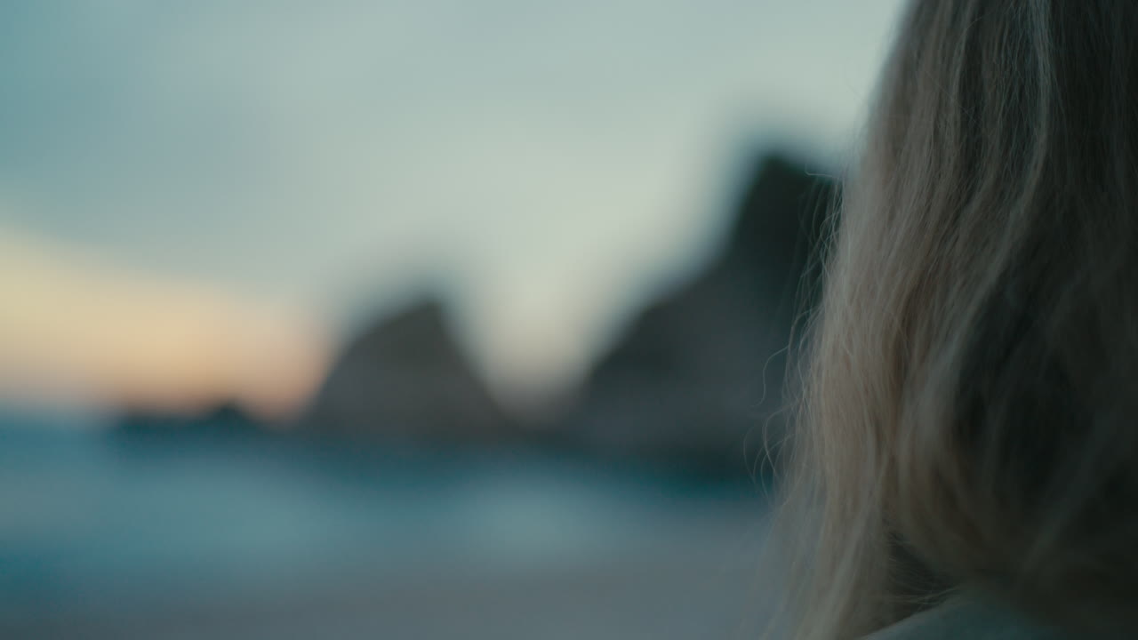 Primo piano sognante con profondità di campo ridotta, con i capelli biondi della donna a fuoco e la costa rocciosa e l'oceano sfocati sullo sfondo al tramonto.