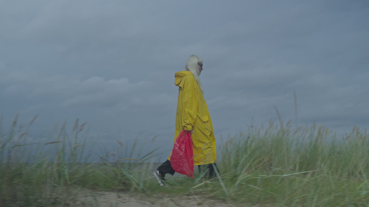 mujer en impermeable amarillo con bolsa roja caminando a lo largo de la duna de arena, capturada con una pista de cámara lateral