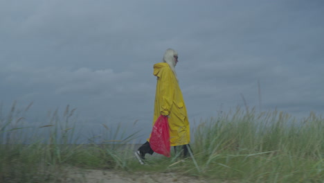 Mujer-Con-Impermeable-Amarillo-Y-Bolsa-Roja-Caminando-Por-Una-Duna-De-Arena,-Capturada-Con-Una-Cámara-Lateral