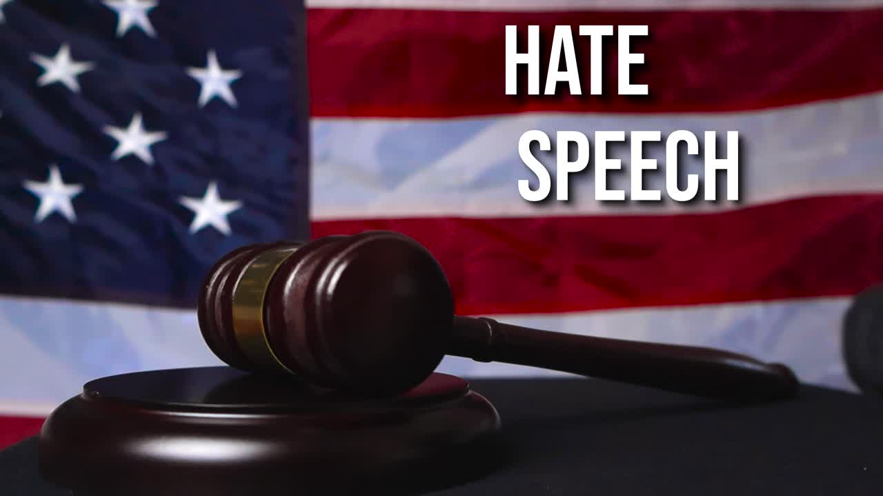 Close up view of wooden brown legal justice court hammer slamming twice on a wooden table and the words Hate Speech appear in the background on American flag. Hand touching the hammer has black glove
