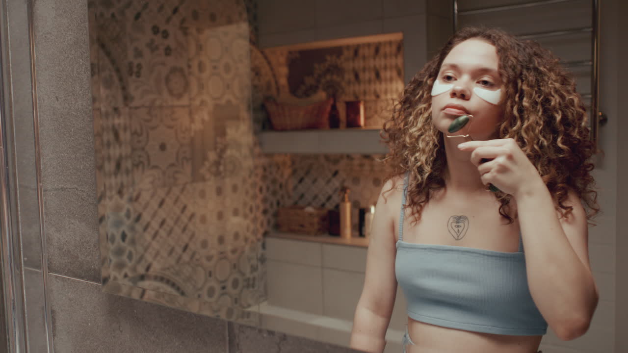Chica con parches debajo de los ojos masajeándose la cara con un rodillo en el baño.