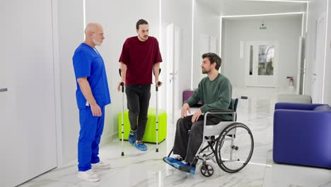 A-confident-male-doctor-with-a-gray-beard-and-glasses-in-a-blue-uniform-communicates-with-two-guys-after-injuries-during-rehabilitation-with-crutches-and-a-special-chair-for-the-disabled-in-a-modern-clinic