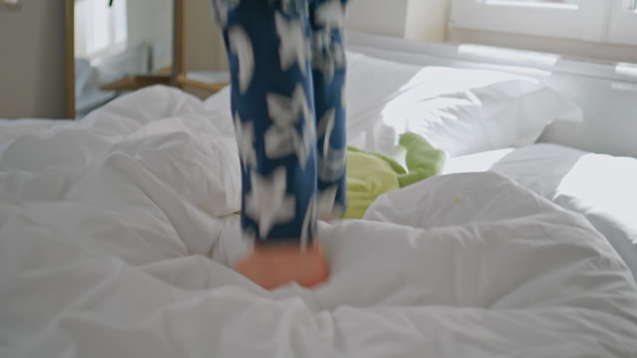 Overjoyed toddler jumping bed having fun with unrecognizable mom at home closeup