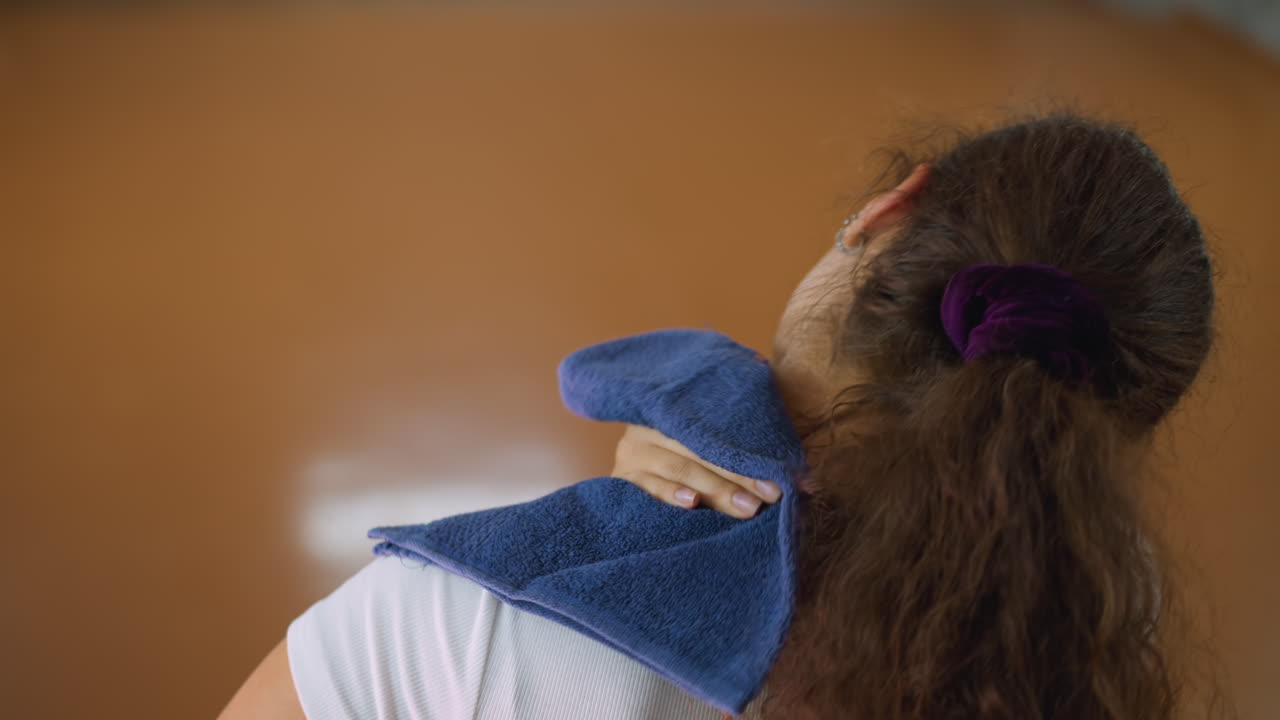 Back view of woman with long curly hair tied in scrunchie wiping sweat from neck using towel after workout in gym environment showing fatigue recovery effort and determination