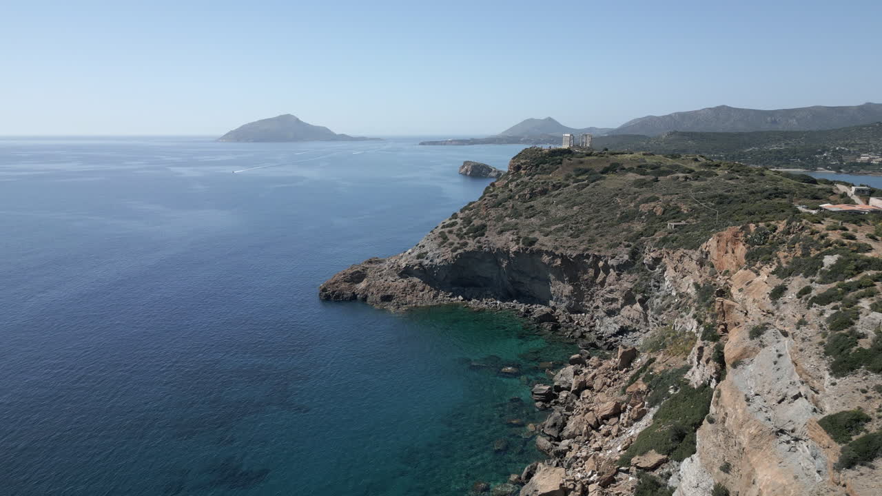 A drone captures the breathtaking view along a cliff in Sunion, Greece, ending with the ancient ruins of the Temple of Poseidon, with an island visible in the distance