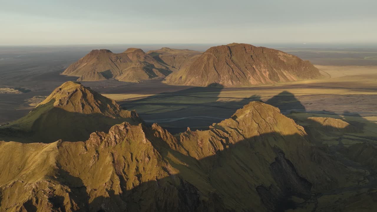 An aerial view of Thakgil, Iceland, showcasing rugged volcanic peaks and expansive shadowed plains bathed in soft golden light.