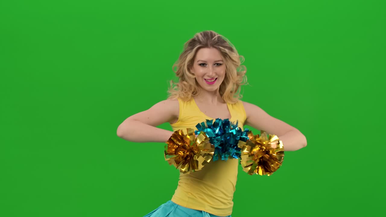 Rehearsal of a dance of a cheerleader in a yellow-blue uniform on a green screen in a studio. A young cheerleader dances an uplifting dance, moving her arms and hips vigorously. Close up. Slow motion