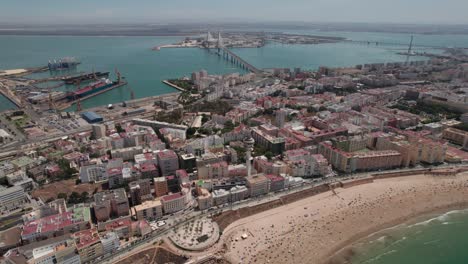 Drone-Aéreo-Hacia-Adelante-E-Inclinado-Hacia-Arriba-De-La-Ciudad-De-Cádiz-Con-Vista-Al-Puente-De-La-Constitución,-Zona-Costera-Del-Puerto,-Cádiz-España