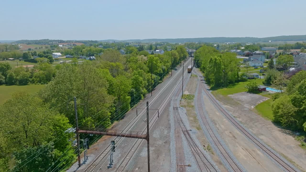 The serene landscape features intersecting train tracks surrounded by vibrant green trees and open fields. A few trains pass near the junction, of a new freight yard, creating a lively atmosphere.