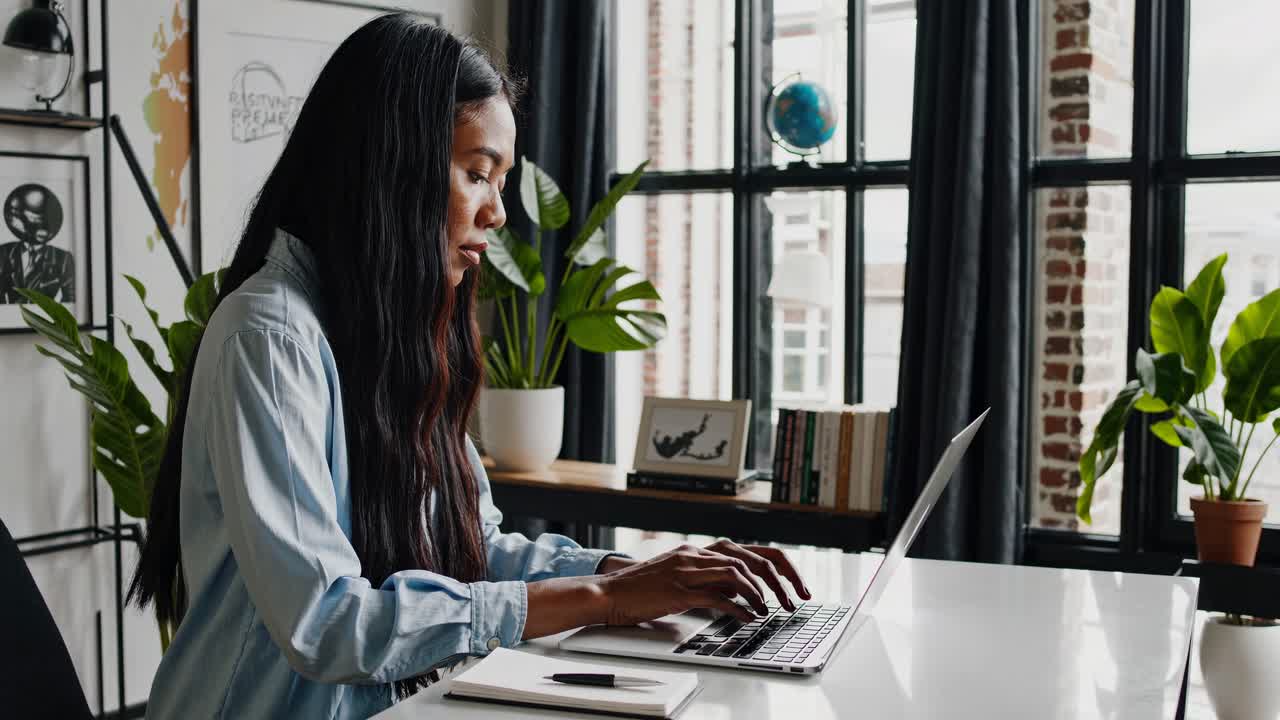 A woman working on a laptop at a desk in a modern home office. Side angle captures a video call