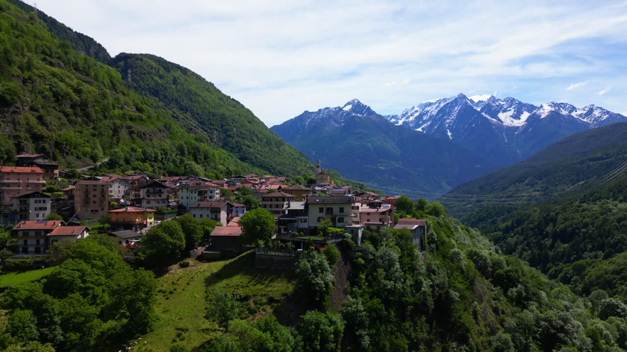 Aerial shot of Malonno village perched on a forested hillside with snowy Alpine peaks in the distance. Shot at Malonno, Italy (Italia)