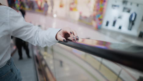 woman with black polished nails gently tapping escalator handrail while ascending, she wears a white blouse and jeans, soft-focus background captures a modern shopping mall with blurred shoppers