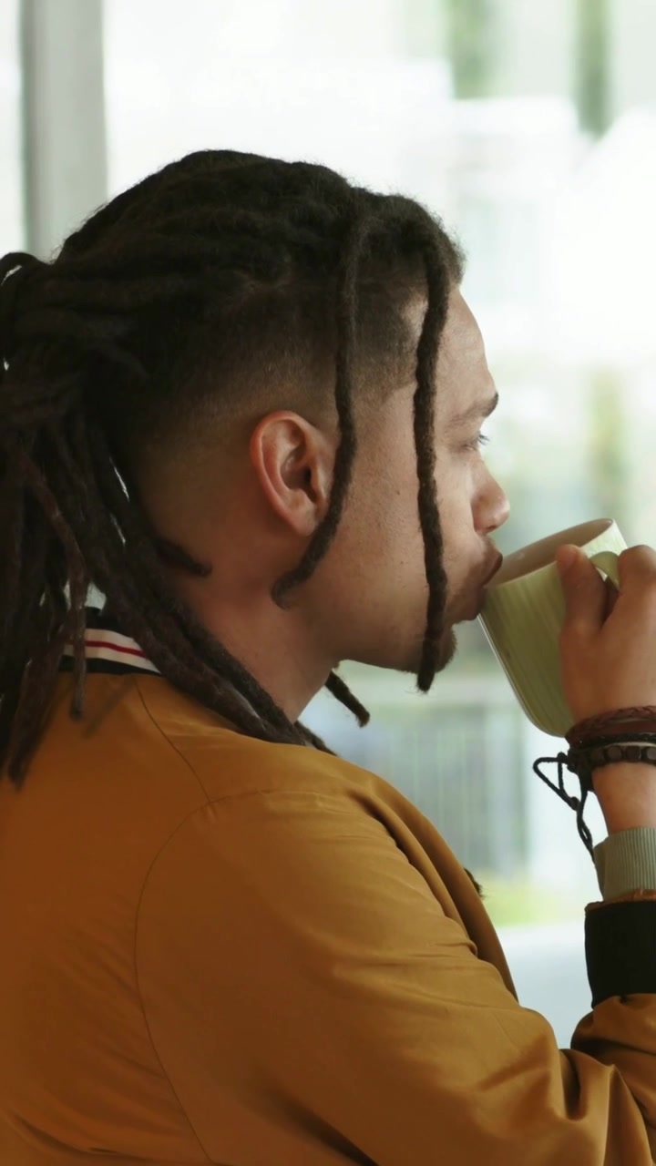 Vídeo vertical: Homem com dreads olhando de relance, bebendo de caneca verde perto da janela, com jaqueta mostarda e pulseiras