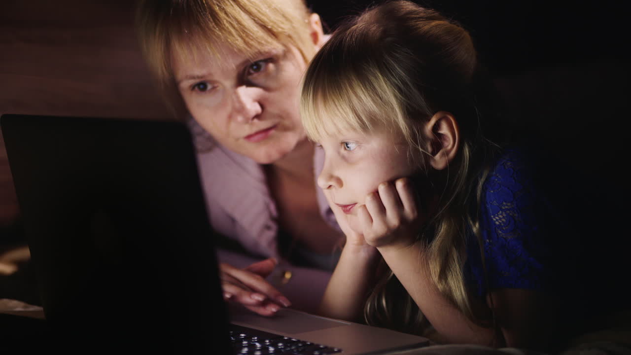 딸과 함께 젊은 어머니가 침대 옆에 누워 있는 노트북에서 함께 놀고 있습니다.