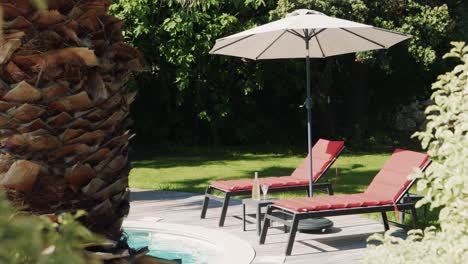 two-empty-red-loungers-by-a-pool-with-a-white-umbrella-in-a-luxurious-garden-with-palm-tree-trunk-and-lawn