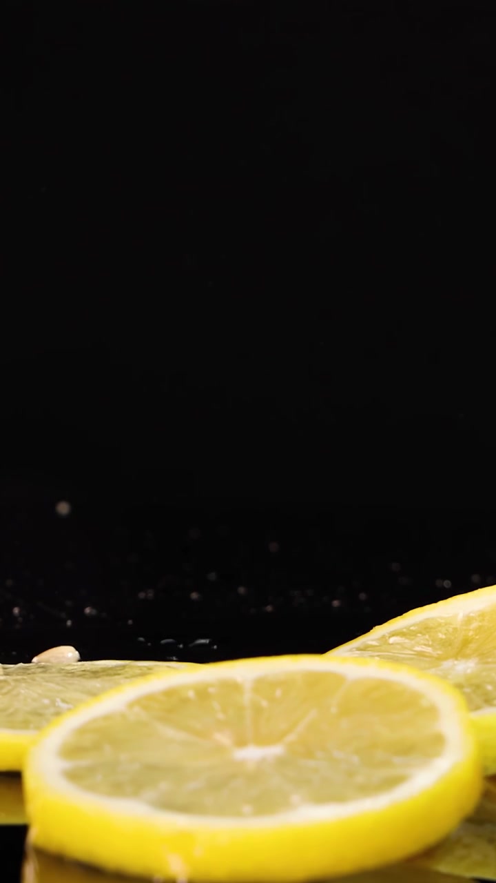 Sottili fette di limone cadono e atterrano su una superficie nera lucida, schizzando goccioline in un'illuminazione da studio drammatica con un'angolazione della telecamera ravvicinata e ad alta velocità.