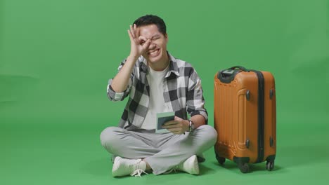 full body of asian male traveler with luggage and passport smiling and showing ok hand sign over eye while sitting in the green screen background studio