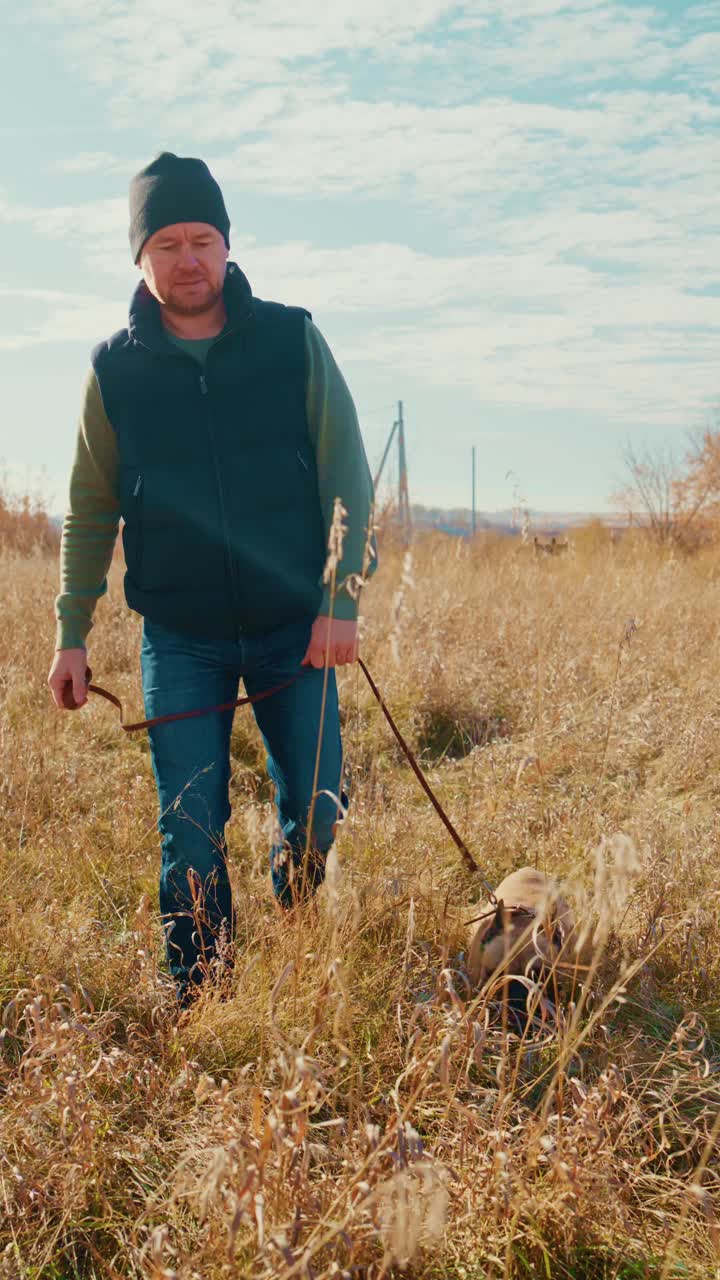 Um homem passeando com seu cachorro por um campo aberto em um dia claro, cercado por gramíneas altas e uma paisagem serena, personificando a beleza tranquila da natureza e a alegria da companhia