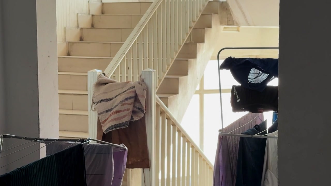 A static shot looking through a doorway at wet clothes hanging on drying racks inside an apartment building stairwell. Bright daylight streams through the background window illuminating the stairs.