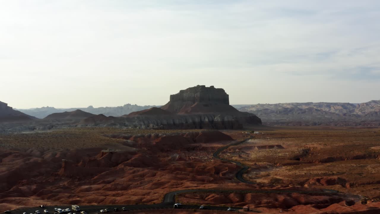 Una scena desertica aerea ripresa da drone in ascesa, con una strada stretta e tortuosa circondata da rocce rosse e sabbia e grandi formazioni rocciose a butte rosse e bianche sullo sfondo, presso il Goblin Valley Utah State Park