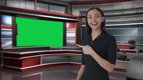 Happy-Indian-female-news-anchor-pointing-at-green-screen-and-showing-thumbs-up