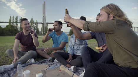 Smiling-young-people-clinking-beer-bottles-and-eating-pizza