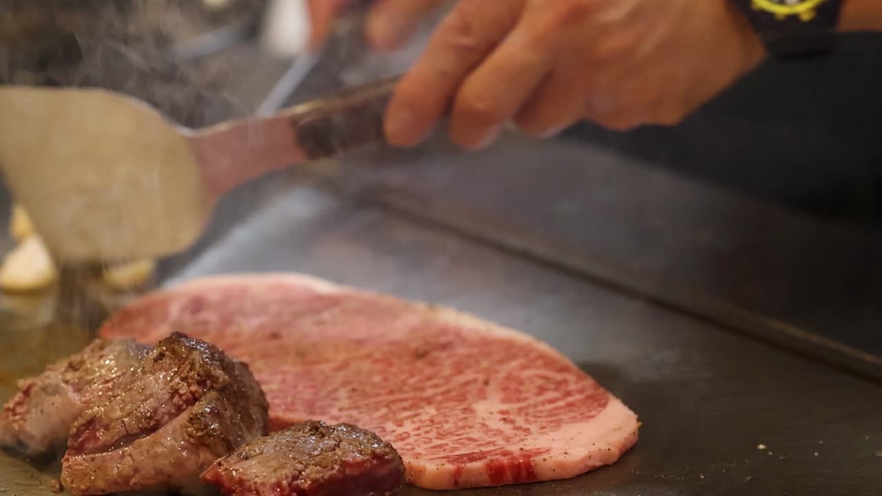 Uno chef usa spatole di metallo per girare e rosolare carne marmorizzata su una piastra calda