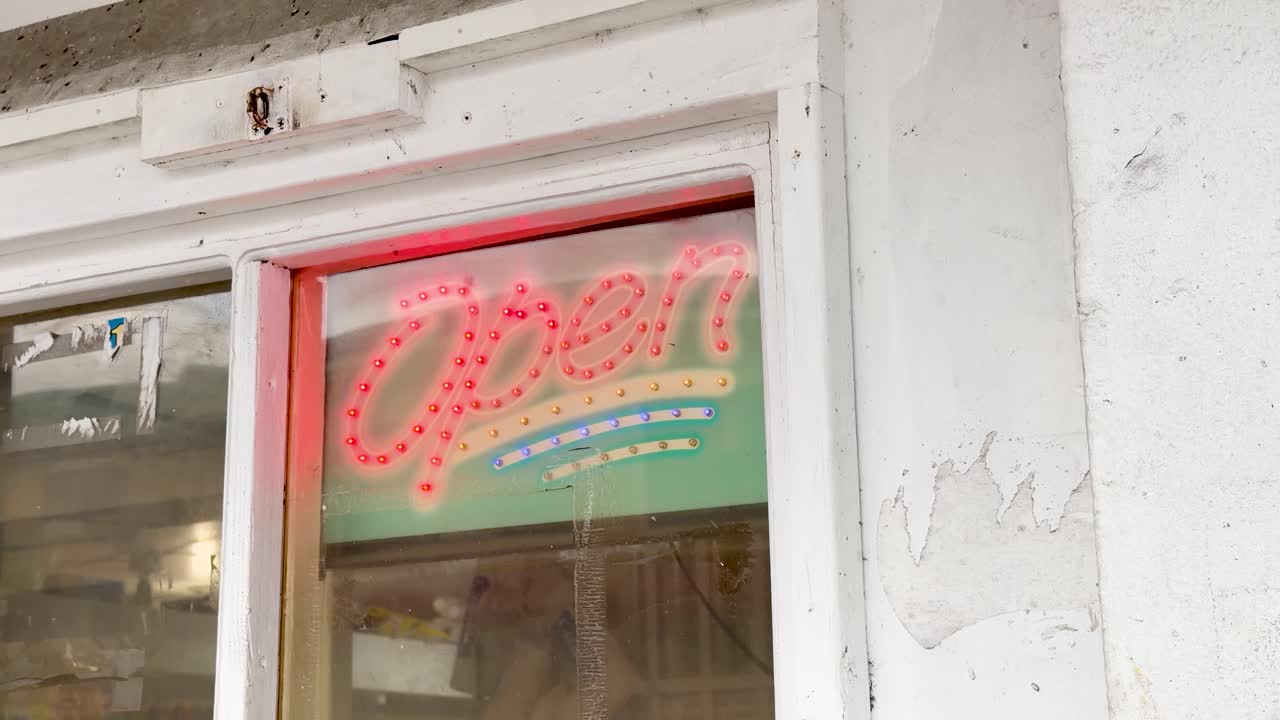 Un'inquadratura media cattura un'insegna al neon "OPEN" tremolante nella vetrina bianca di un negozio al dettaglio.