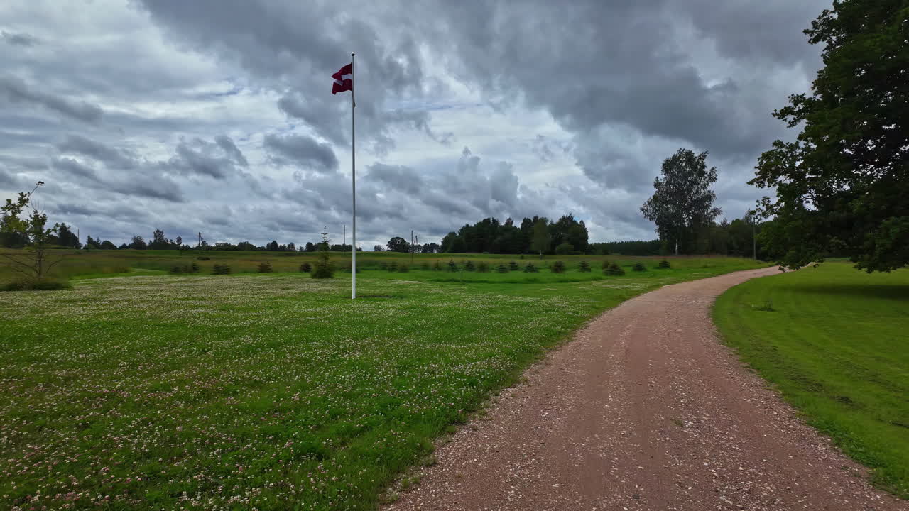 Dirt path winds through a green field with Latvia flag and trees under cloudy skies
