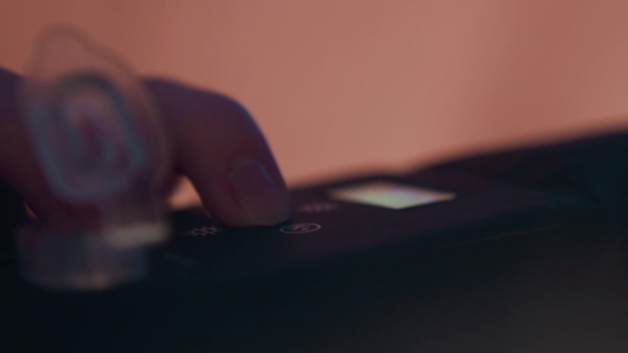 Close-up shows fingertip pressing glowing control on dark device, with slight finger bend, soft blur, warm orange light, and moody contrast emphasizing tactile operation in tight framing