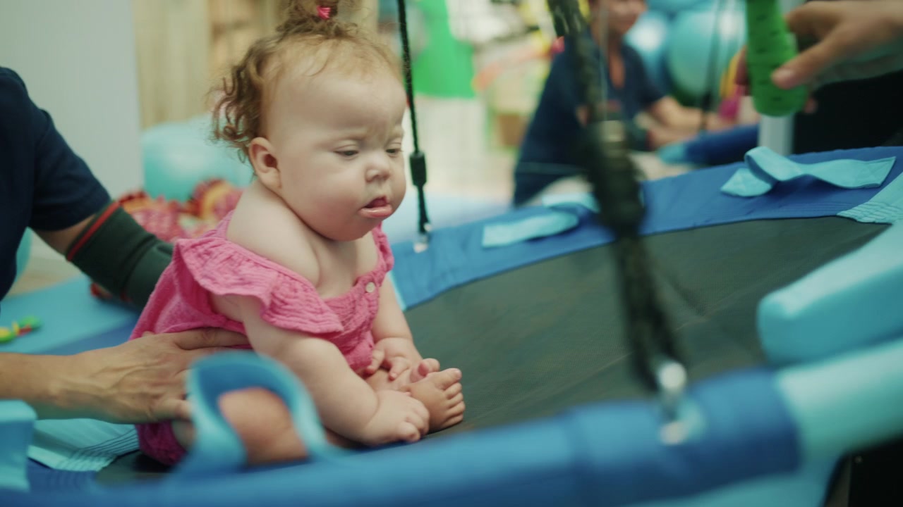 Bebê menina com síndrome de Down sentada em um balanço sensorial, recebendo fisioterapia e reabilitação de um terapeuta.