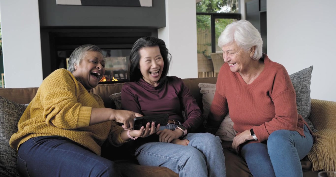 Diverse female friends with senior on couch by fireplace, left woman showing remote, all laughing