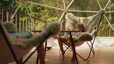 brother and sister sit in authentic fur armchairs in glamping camp while relaxing after walk in forest. children look at mountain scenery outside window