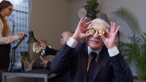 Elderly-man-boss-dancing,-fooling-around,-making-silly-faces-with-bitcoin-coints-eyes-in-office