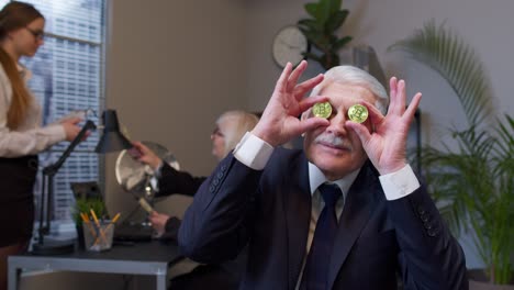 anciano jefe bailando, tonto alrededor, haciendo caras tontas con monedas de bitcoin ojos en la oficina
