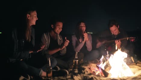 Round-camera-movement:-Multiethnic-group-of-young-people-sitting-by-the-bonfire-late-at-night-and-singing-songs-and-playing-guitar,-clapping-hands