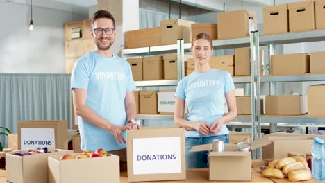 caucasian young male and female volunteers packing box with food and then smiling to the camera in charity warehouse