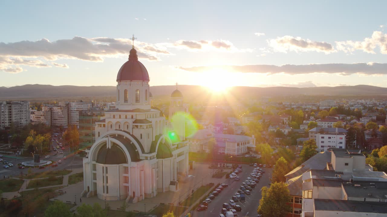 A cinematic aerial shot slowly orbits a majestic Orthodox cathedral in Bacau,Romania. The setting sun creates a spectacular lens flare and bathes the autumn cityscape in warm golden light