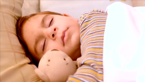 retrato de un niño de 3 años durmiendo con un oso de peluche. la mano de su madre lo cubre con una manta.